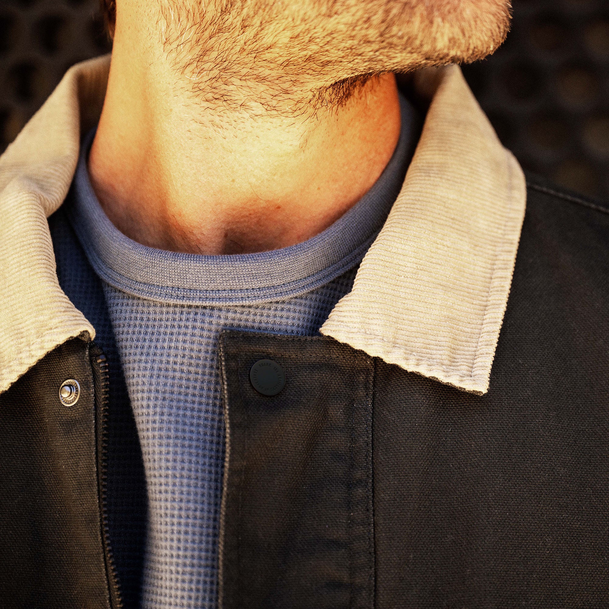 Man wearing black waxed canvas jacket with cream sherpa collar and blue textured shirt underneath
