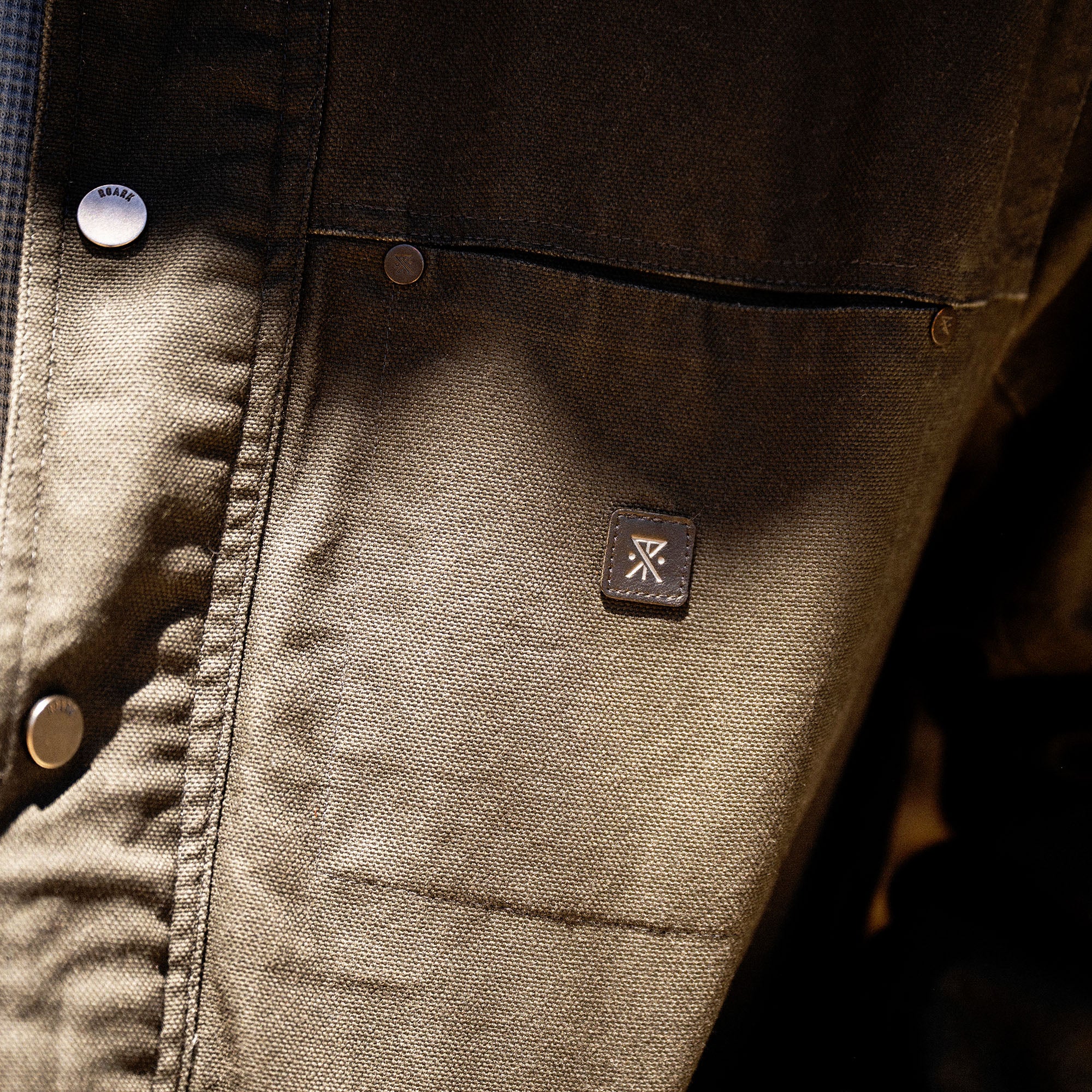 Olive waxed canvas jacket detail showing branded metal button and embroidered logo patch on front pocket