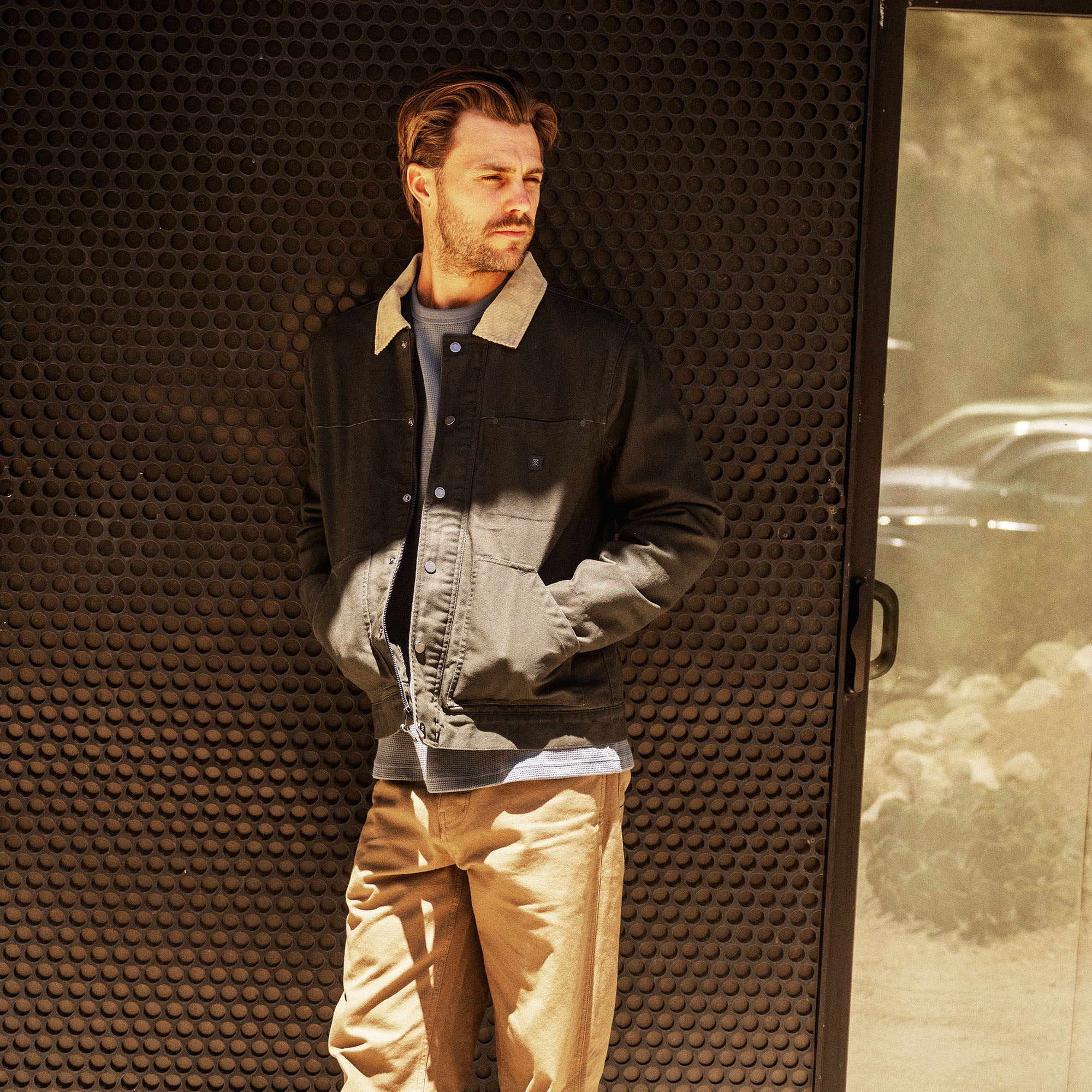 Man wearing black and gray waxed canvas jacket with sherpa collar and tan pants against perforated metal backdrop