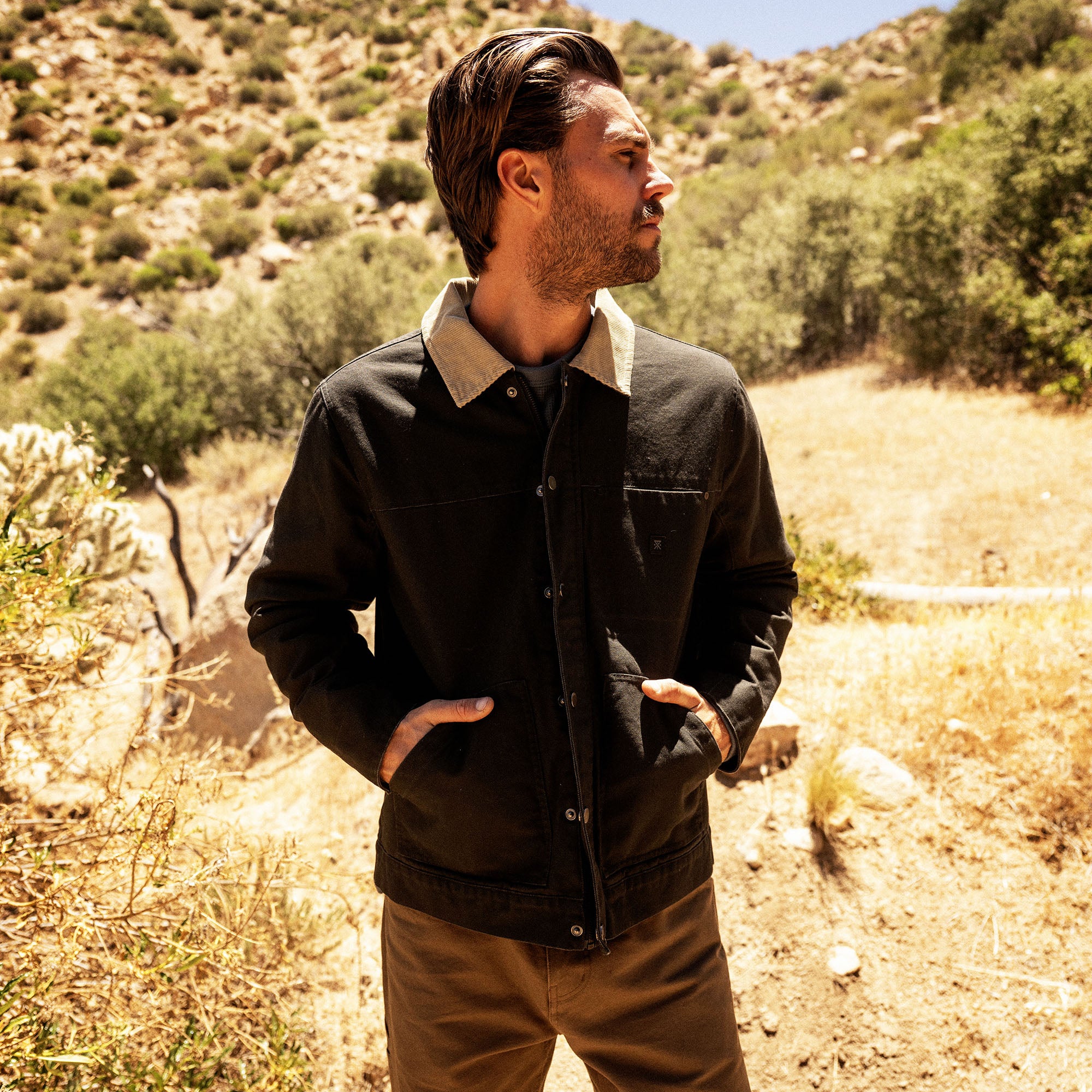 Man wearing black waxed canvas jacket with tan collar, standing on desert trail surrounded by shrubland and rocky hills