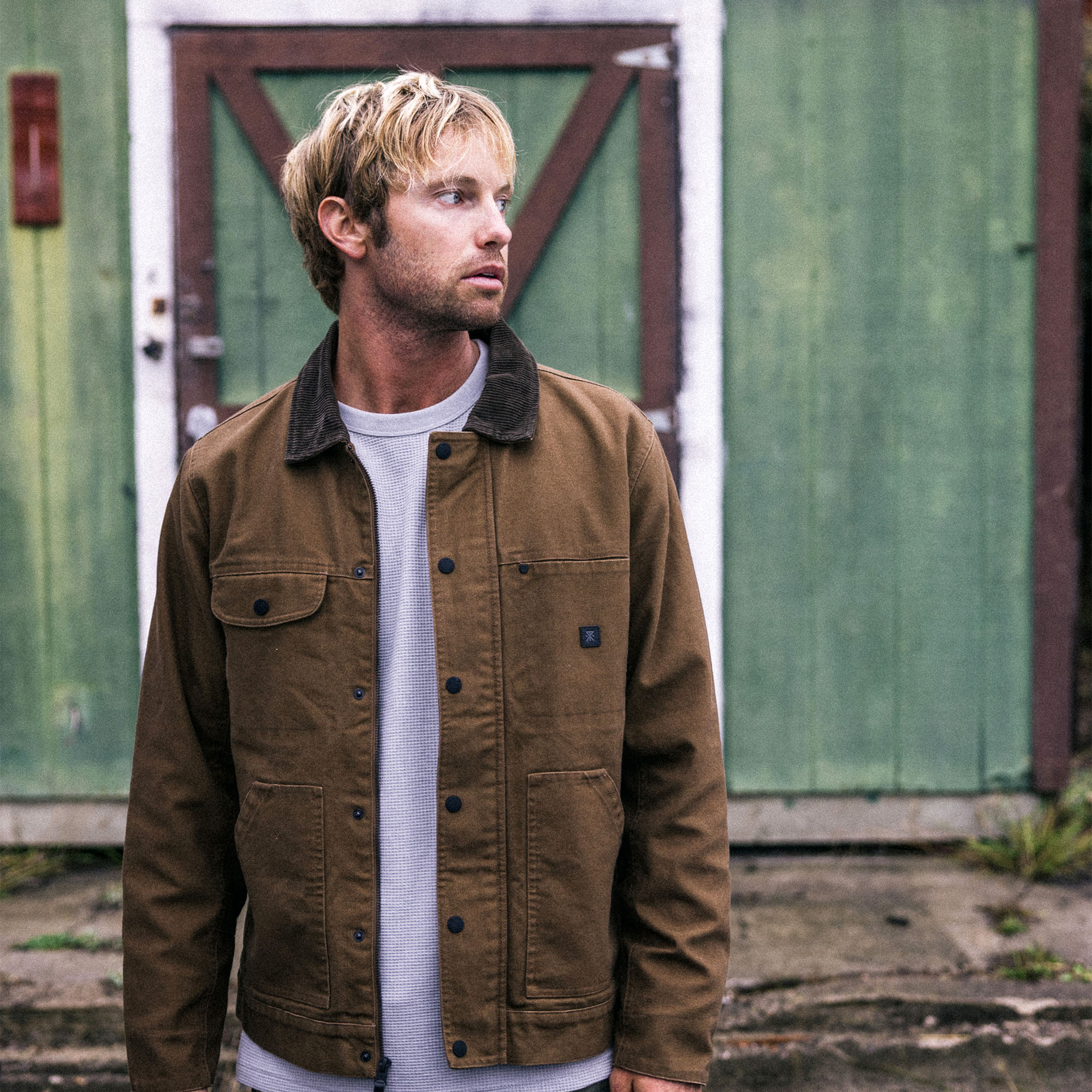 Roark's Open Roads Waxed Canvas Jacket set against a scenic backdrop with a quaint cottage and rustic barn, evoking the spirit of adventure and road trips.