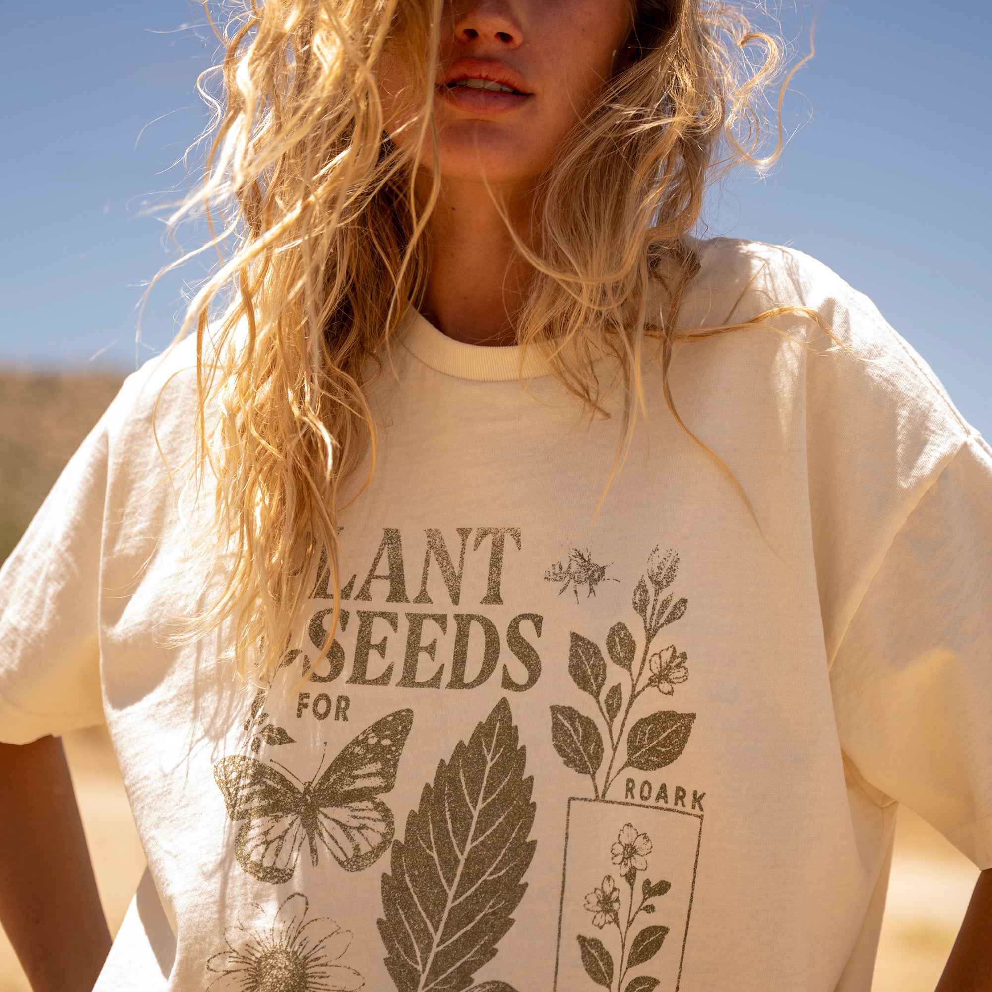 Cream-colored oversized tee with "Plant Seeds for Roark" text and botanical illustrations including butterfly, leaves, and flowers