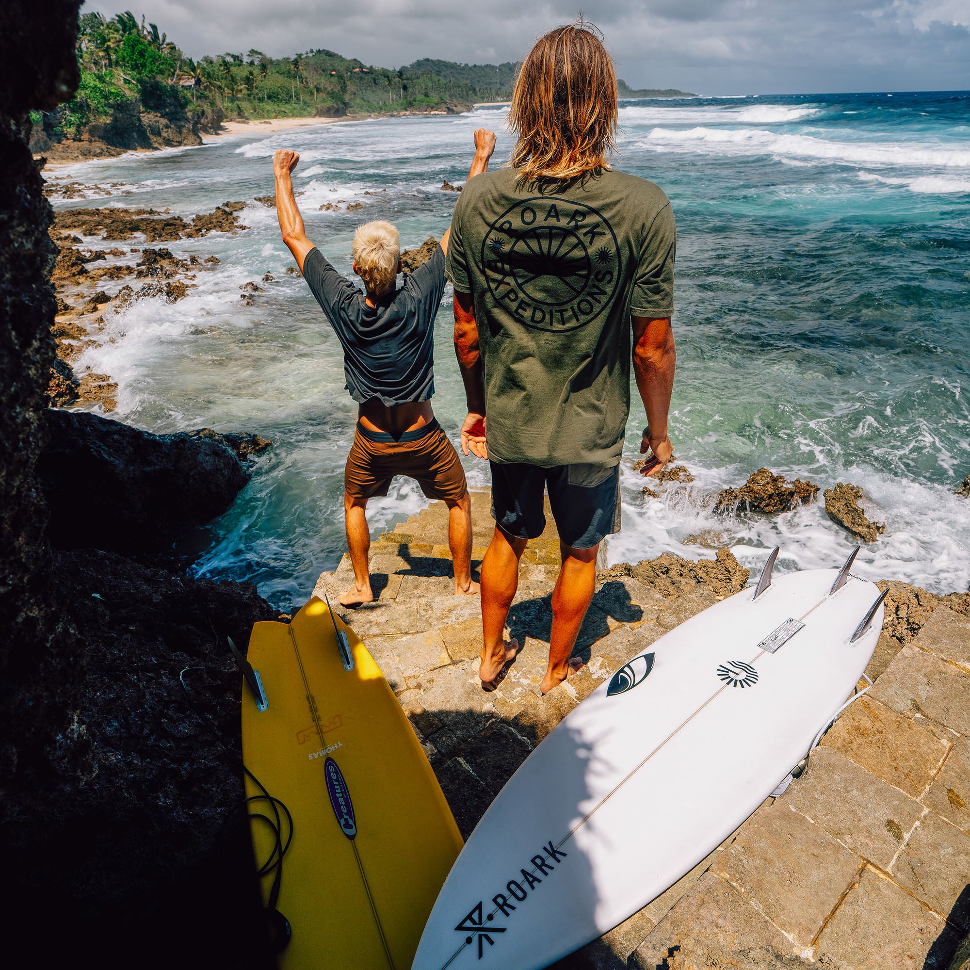 Two surfers in wetsuits standing on rocky beach overlooking tropical ocean waves with surfboards laid out on sand