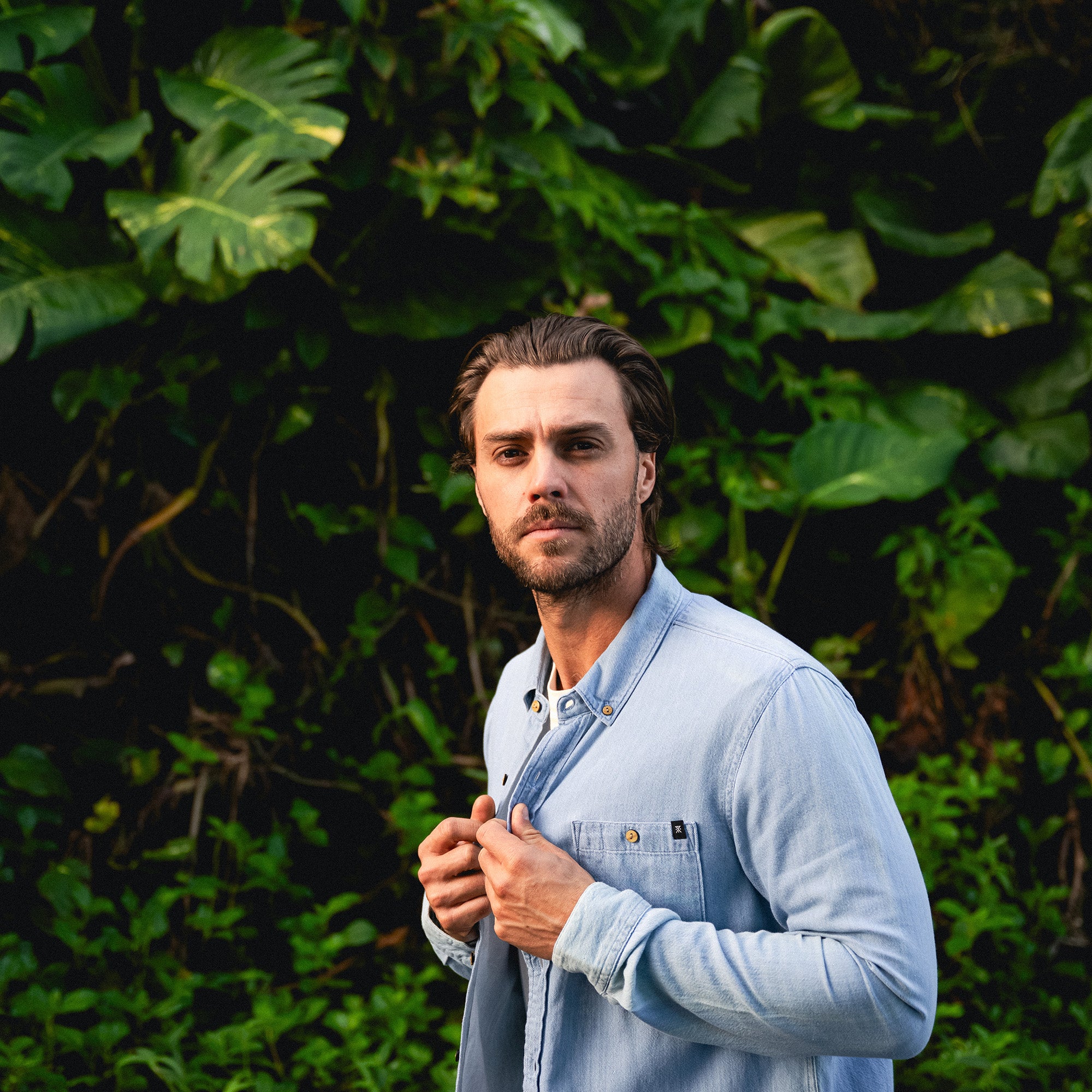 Man with a beard modeling the Scholar Chambray Button Up Shirt featuring detailed sleeves and buttons, perfect for adventure and travel.