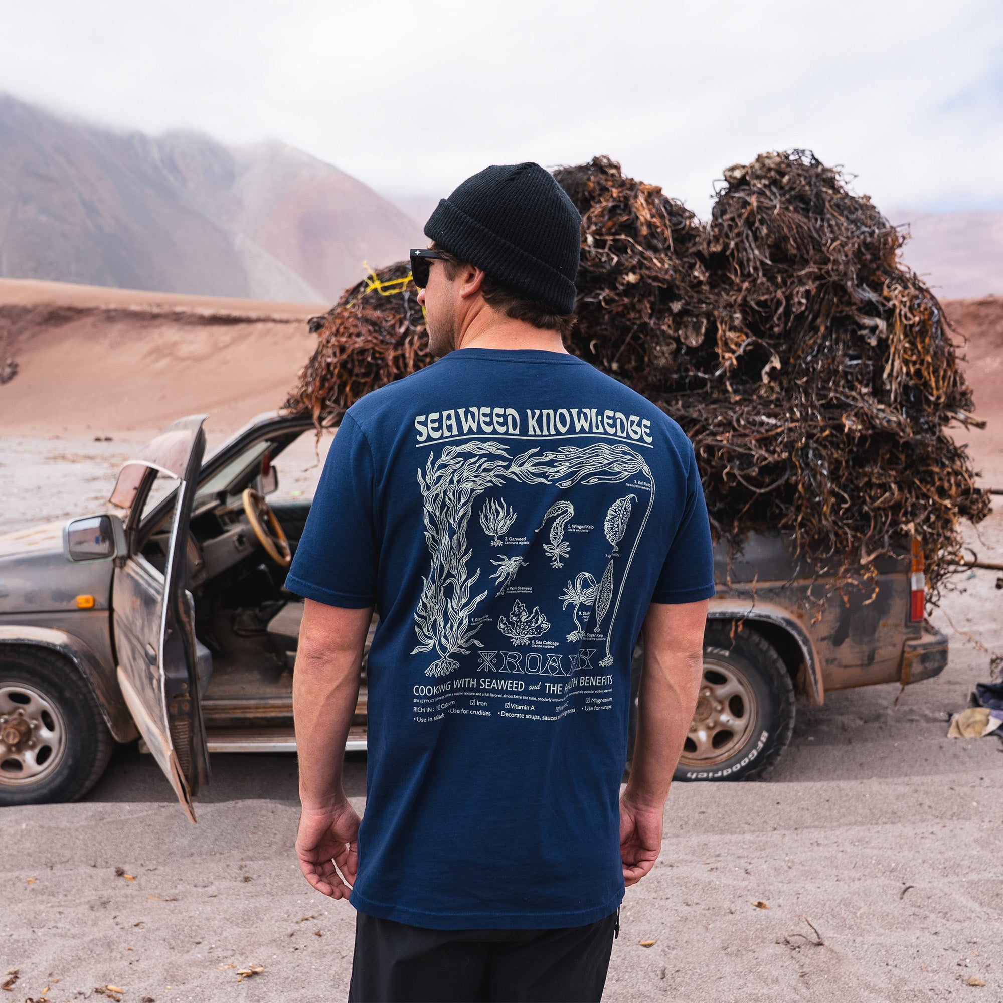 Active man wearing the Roark Seaweed Knowledge Premium Tee and a baseball cap on a beach vacation, epitomizing adventure and outdoor lifestyle.