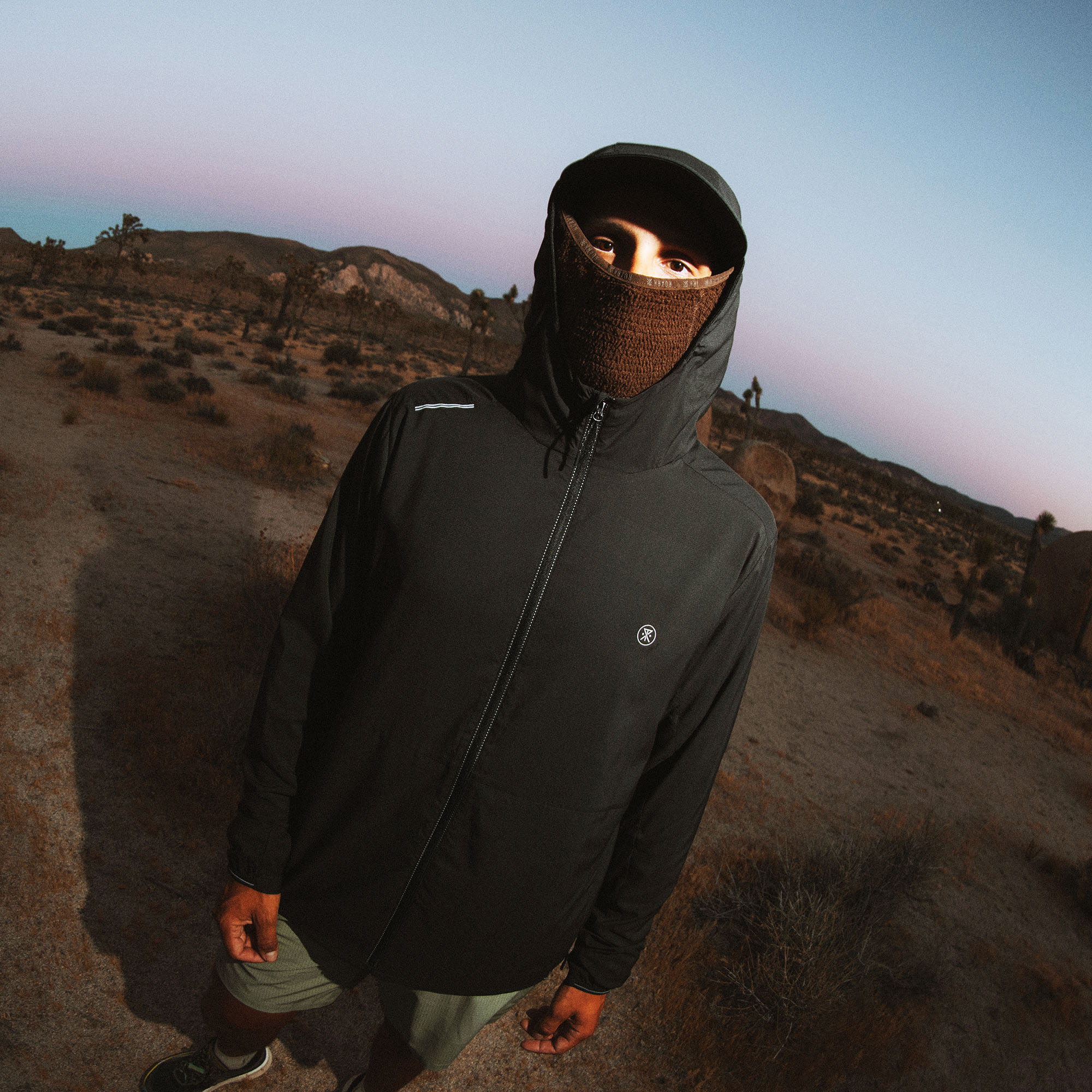Person wearing black Secondwind 3.0 jacket with brown knit face mask in desert landscape at dusk