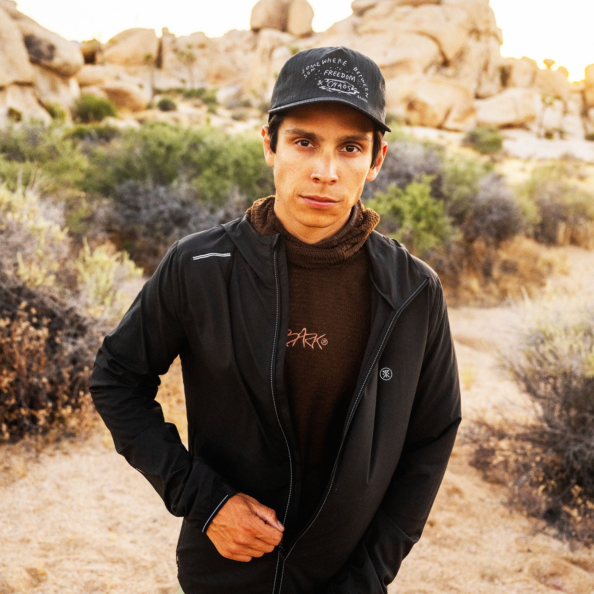 Man wearing black zip-up jacket with brown turtleneck and black baseball cap in desert landscape with boulders