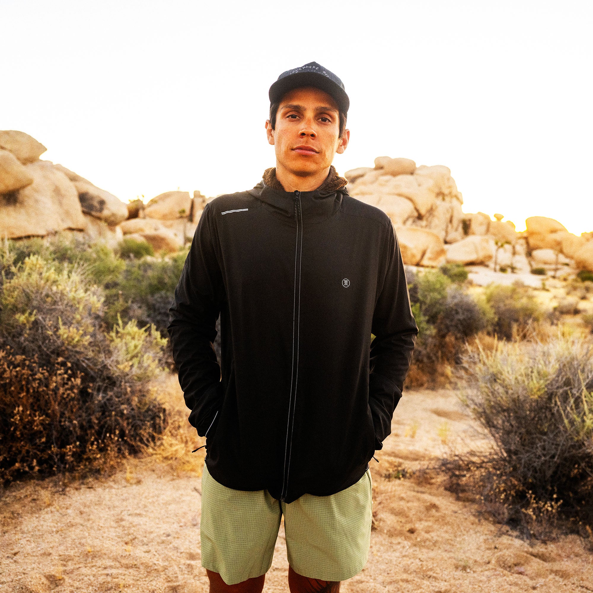 Man wearing black zip-up hoodie jacket with small logo in desert landscape with boulder formations and shrubs