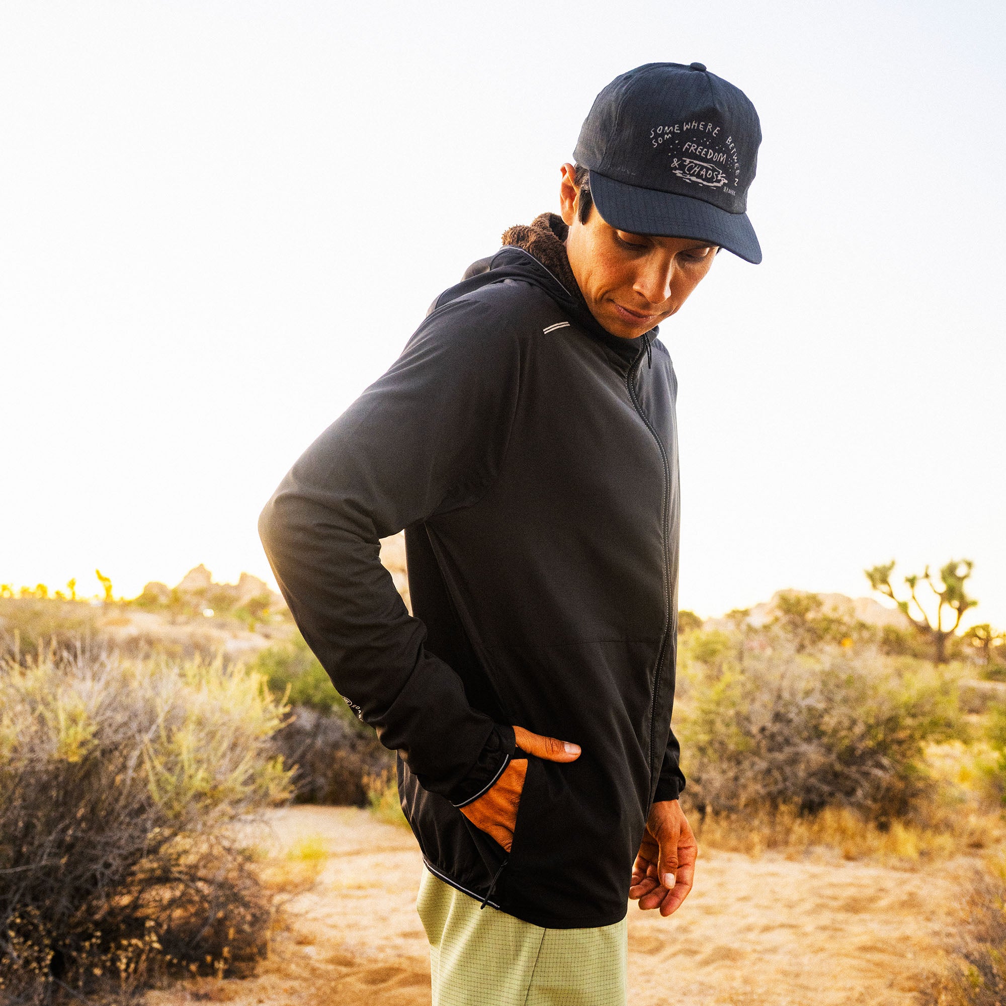 Person wearing black Secondwind 3.0 jacket with orange accents and navy cap in desert landscape at sunset