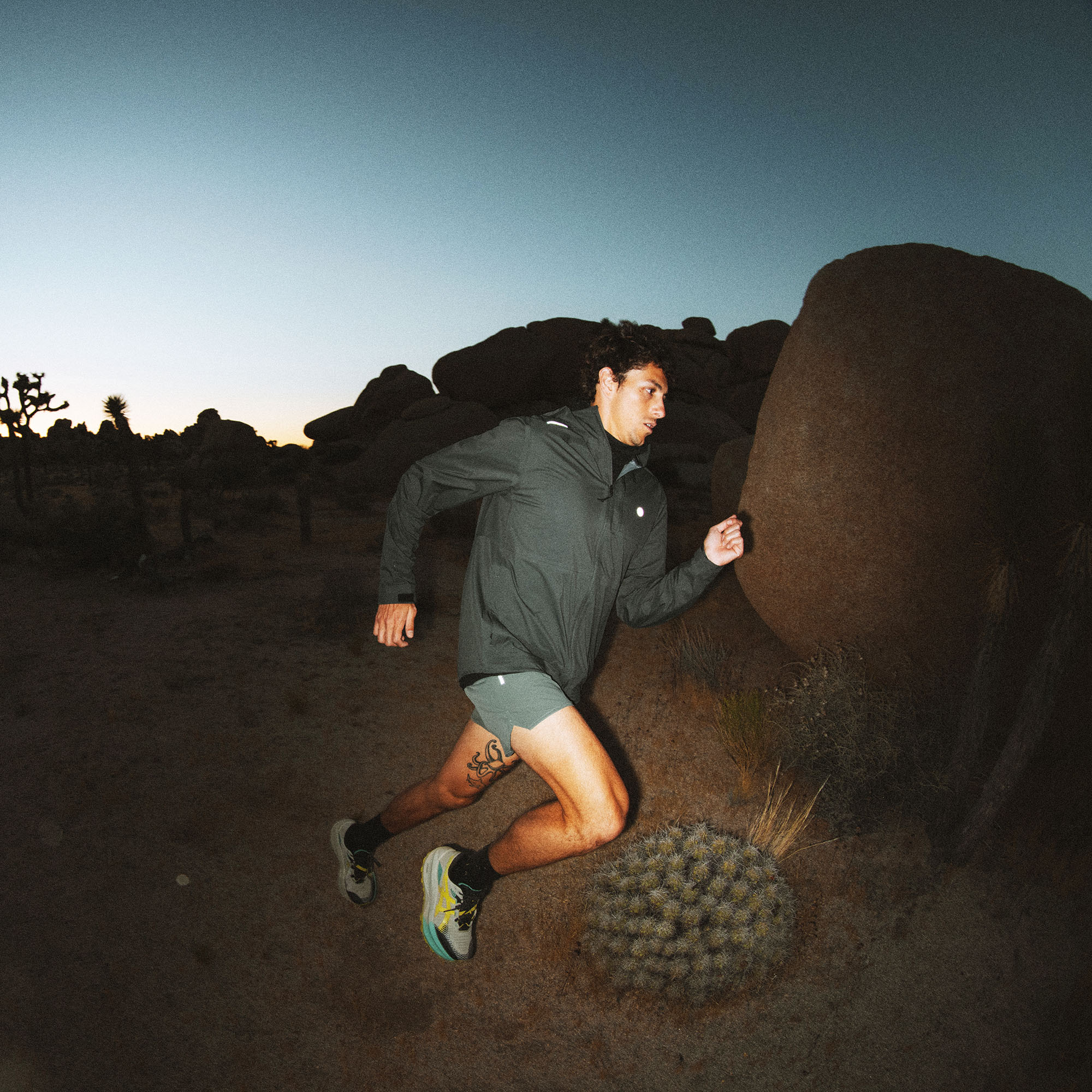 Man running in dark charcoal Shield Rain Jacket near large boulders in desert landscape at dusk