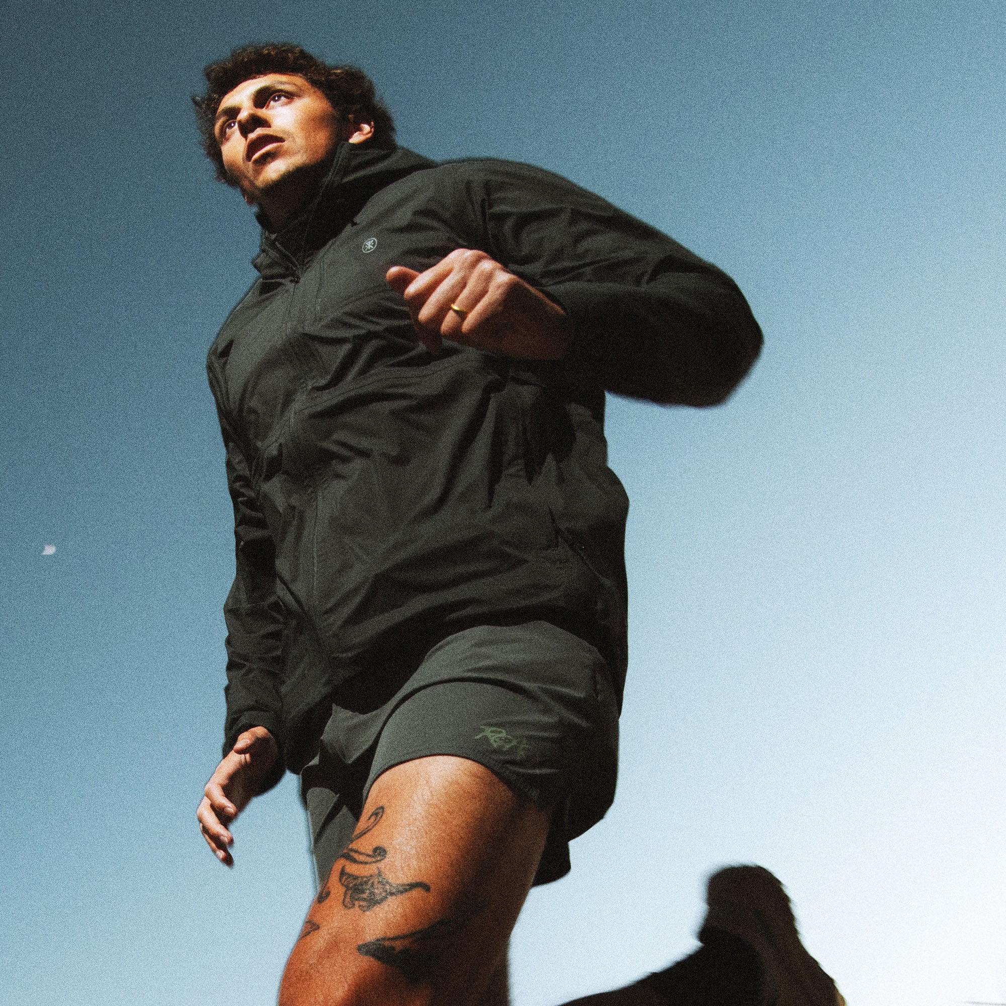 Man wearing black Shield Rain Jacket with shorts, photographed outdoors against clear blue sky in lifestyle shot