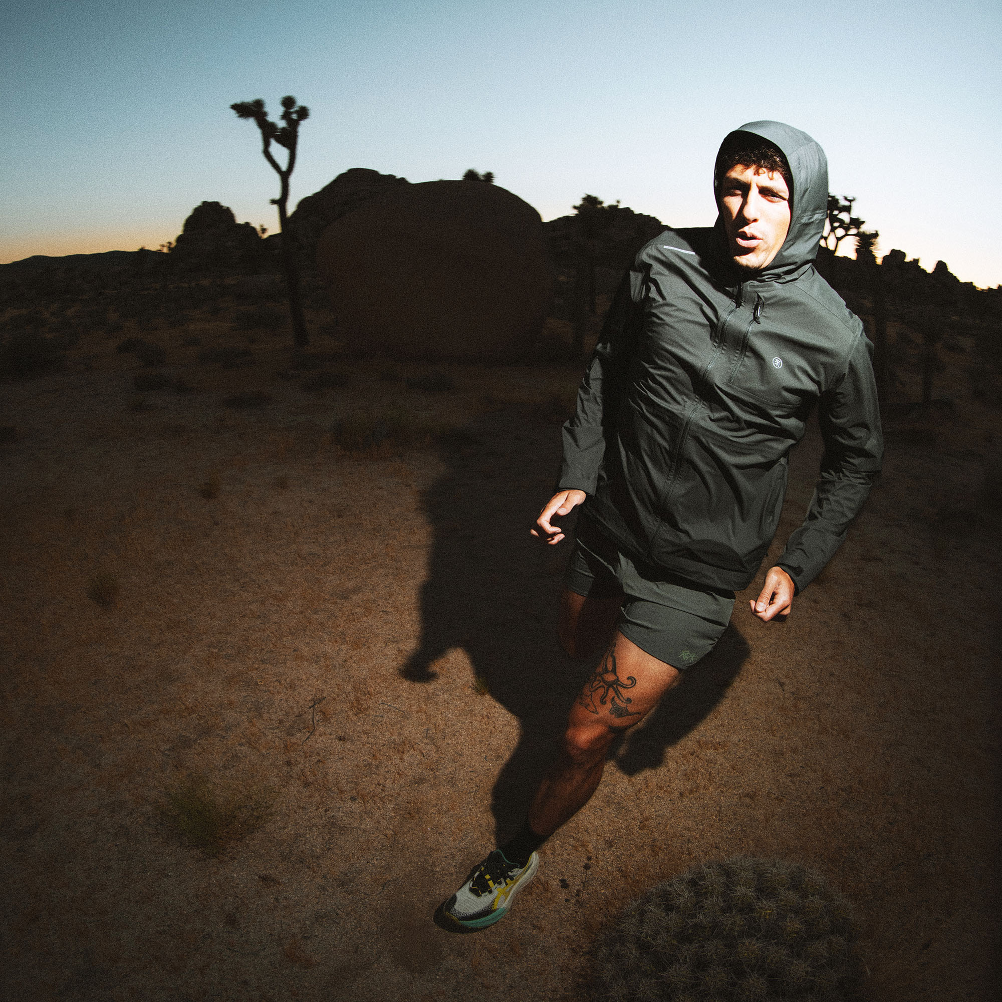 Man jogging in gray hooded rain jacket across desert terrain with Joshua trees at sunset
