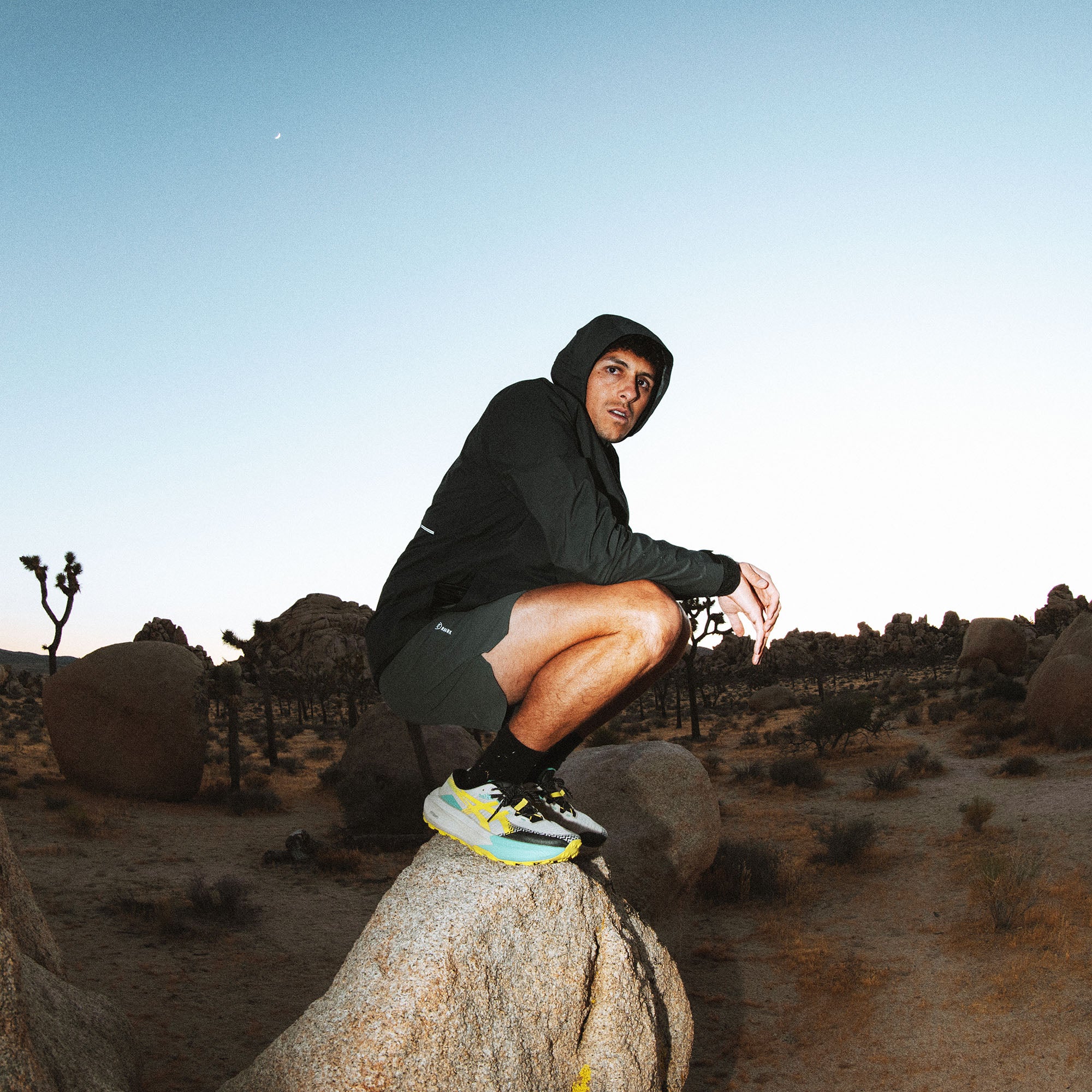 Black Shield Rain Jacket worn by male model crouching on boulder in desert landscape with blue sky