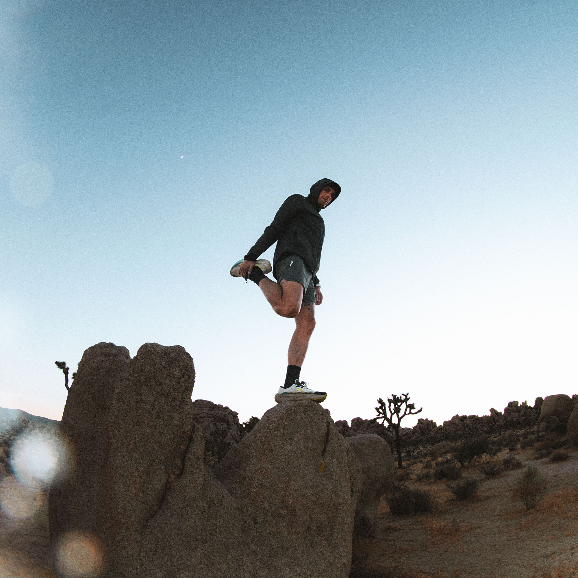 Man wearing black Shield Rain Jacket jogging on desert boulder with clear blue sky and Joshua trees visible in background