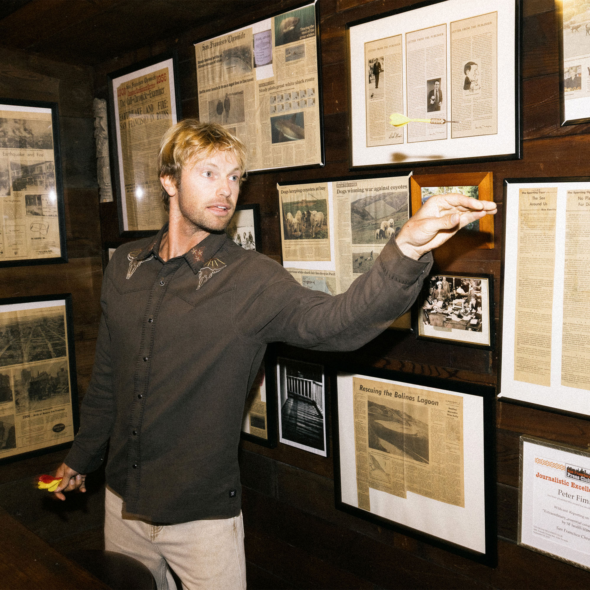 Man with a beard wearing Roark's Tierra del Sur Range Long Sleeve Button-Up Shirt, surrounded by rustic decor and adventure-themed picture frames, embodying a spirit of travel and outdoor exploration.