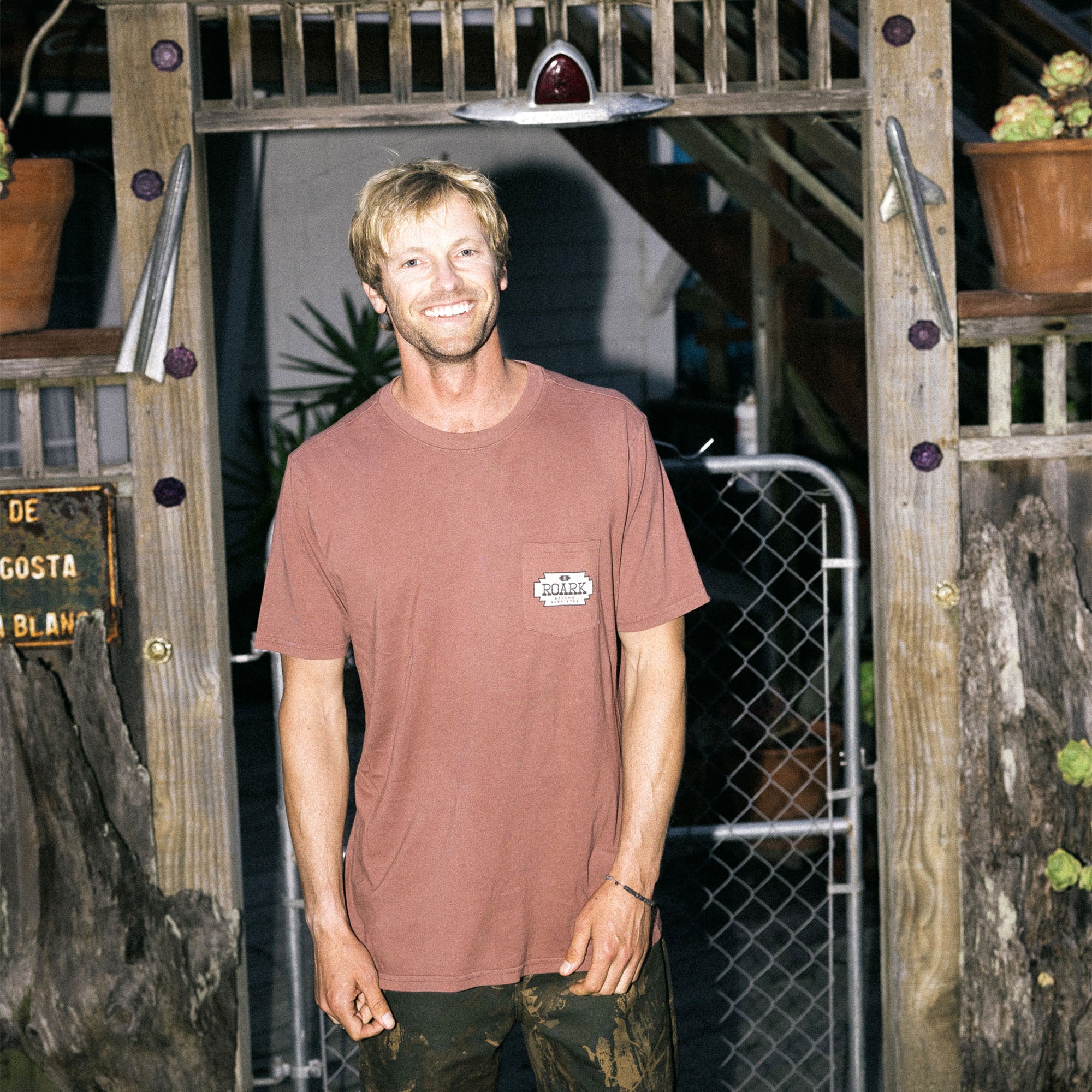 Smiling man with facial hair wearing a Roark Tierra del Sur Shield Pocket Premium Tee, showcasing active outdoor style against a wooden backdrop.