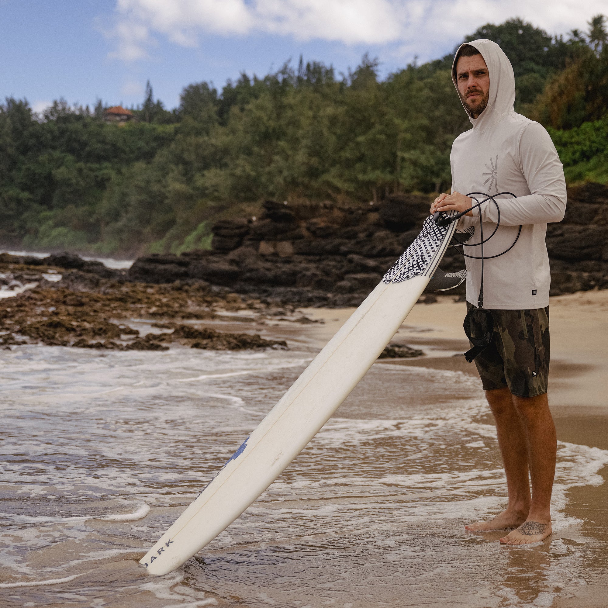 Surfer preparing with a surfboard by the ocean, showcasing the adventurous spirit ideal for Roark’s Venture UPF Hoodie.