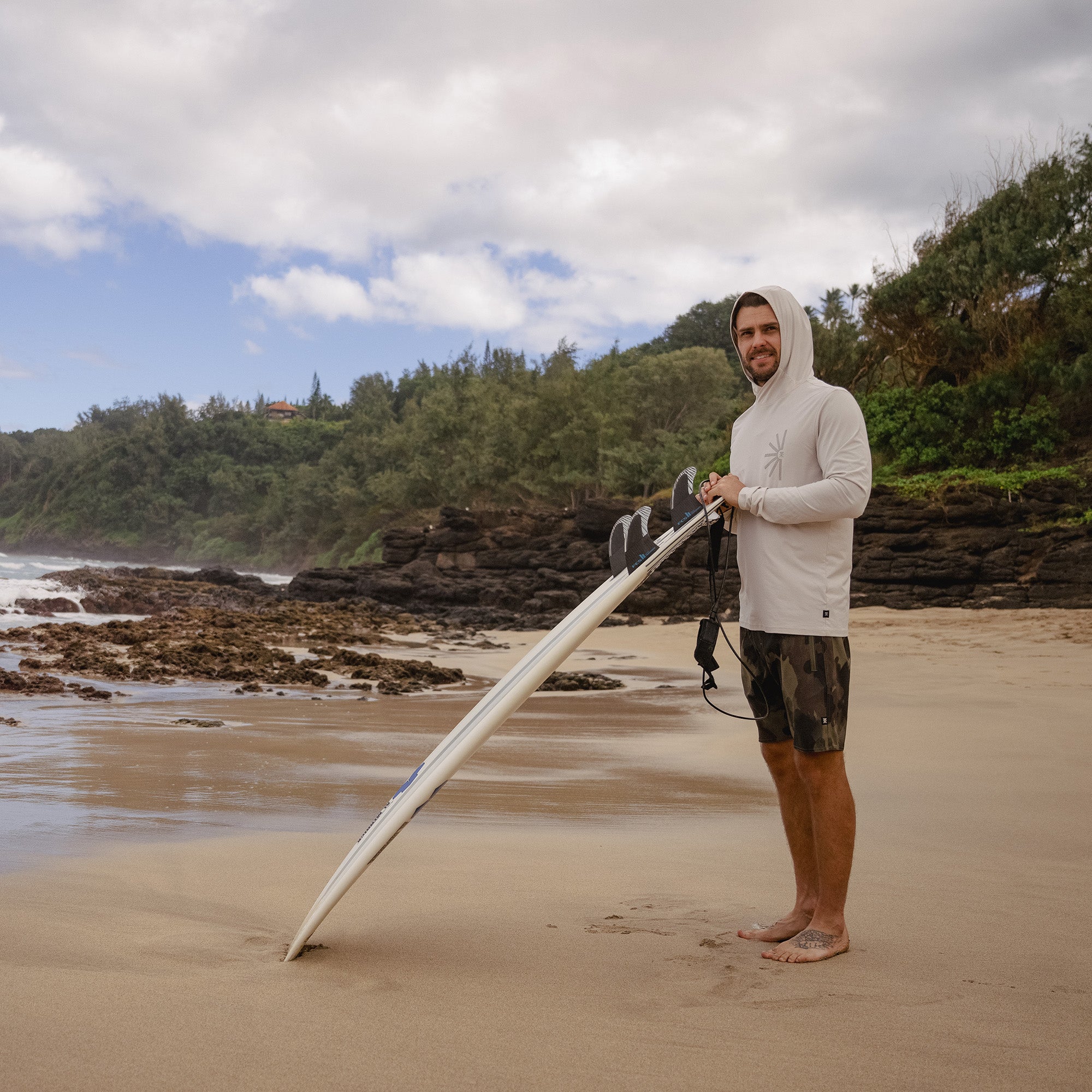 Surfer with a surfboard gearing up in a Roark Venture UPF Hoodie on a sunny beach, showcasing the perfect blend of adventure and sun protection.