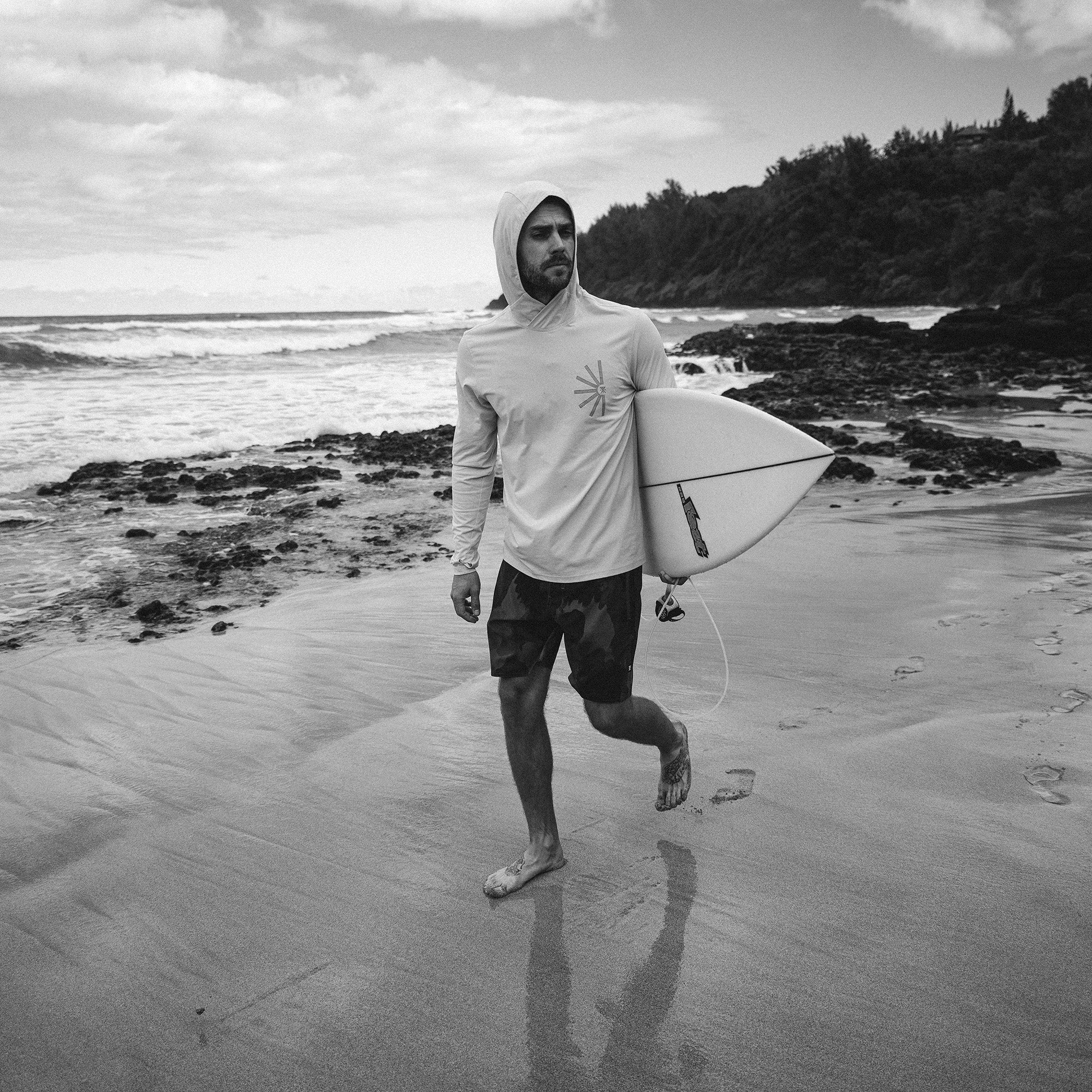 Surfer wearing a Venture UPF Hoodie by Roark, standing on the beach with a surfboard, ready to tackle the waves of the sea, embodying the spirit of adventure and travel.