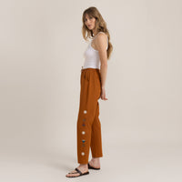 Woman in rust-colored trousers with white tank top and black sandals, side view showcasing casual elegance.